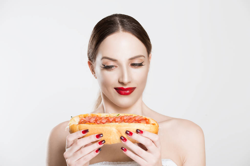 Жажда быстрого питания. Красивая молодая женщина держит в руках вкусный хот-дог, думая съесть его или нет, испытывая сомнения по поводу хот-дог гамбургер изолированный белый фон. Лицо, язык тела
 - Фото, изображение
