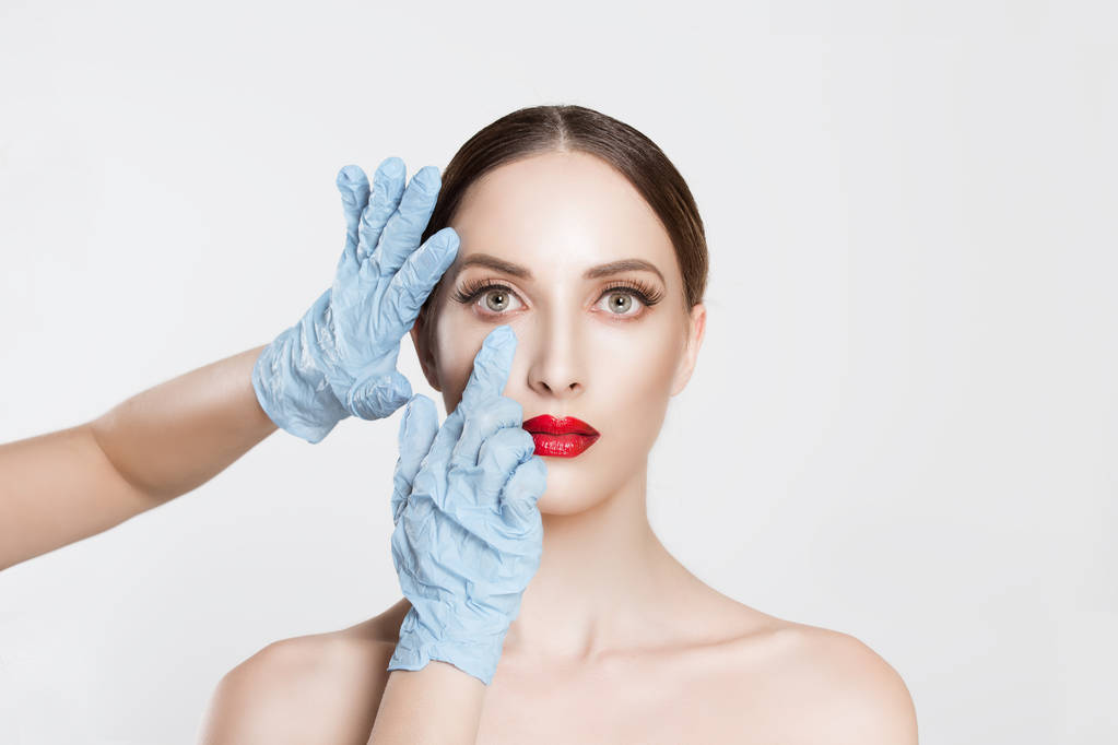 Красота, люди и здоровье. красивая молодая женщина готовится к подтяжке век, блефаропластической хирургии руки врачей в синих перчатках указывая пальцами на ее глаза на белом фоне
. - Фото, изображение