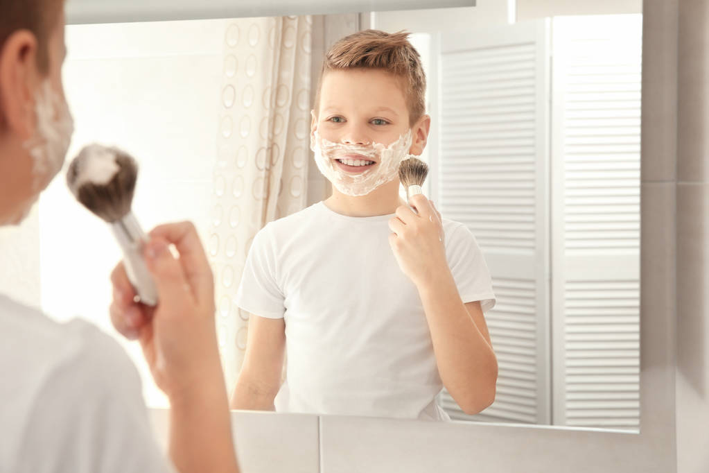 Cute boy with shaving foam on face near mirror in bathroom - Photo, Image