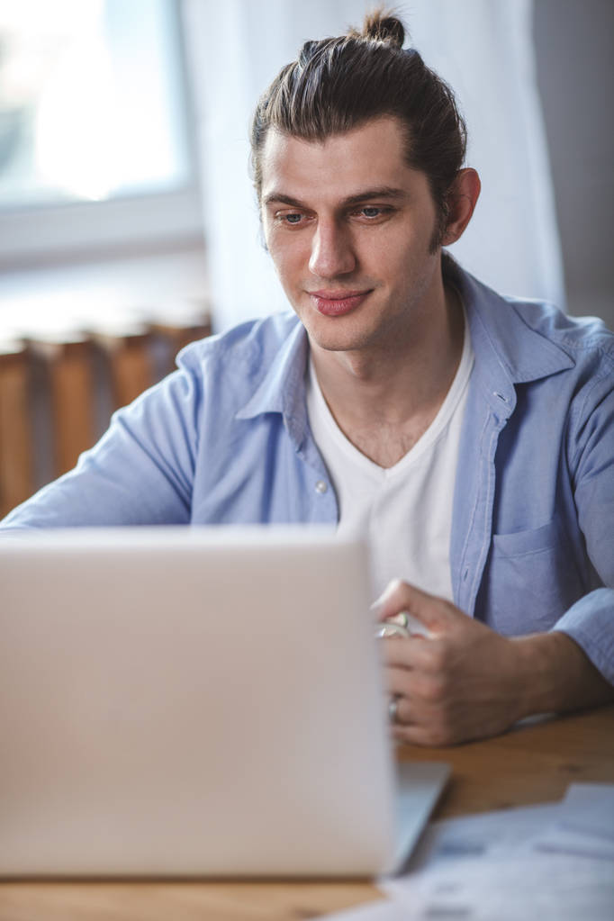 Młody mężczyzna, trzymając kubek, siedzi przy stole i patrząc na ekran laptopa - Zdjęcie, obraz