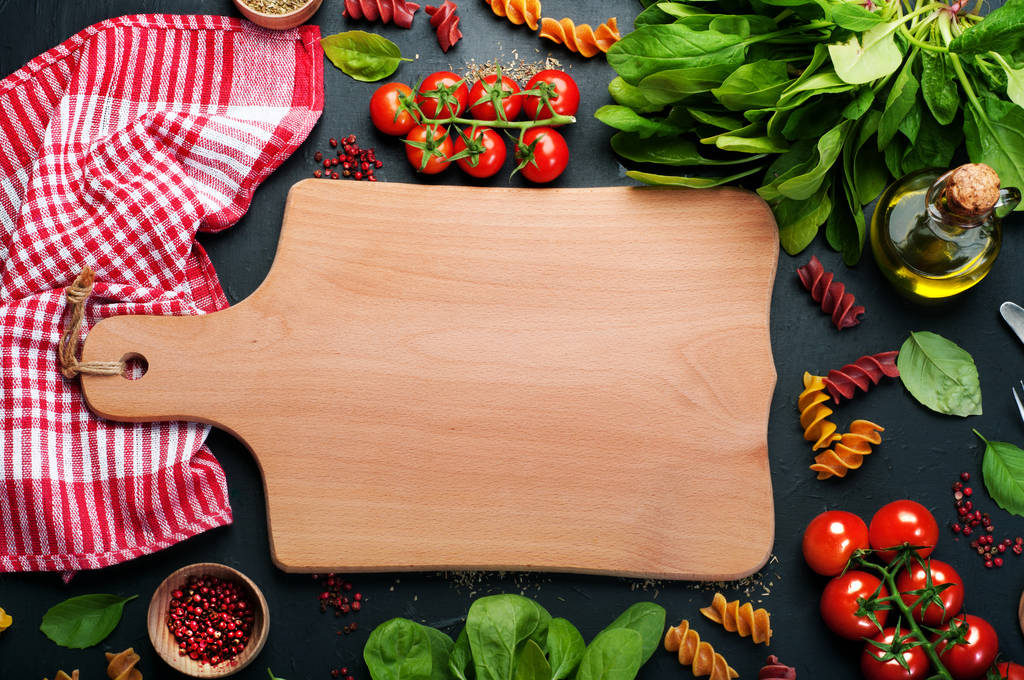 Background. Italian food. Multicolored pasta, tomatoes, spinach, basil leaves and olive oil on a dark surface. In the center of the image is wooden cutting board on which you can arrange dish or text - Photo, Image