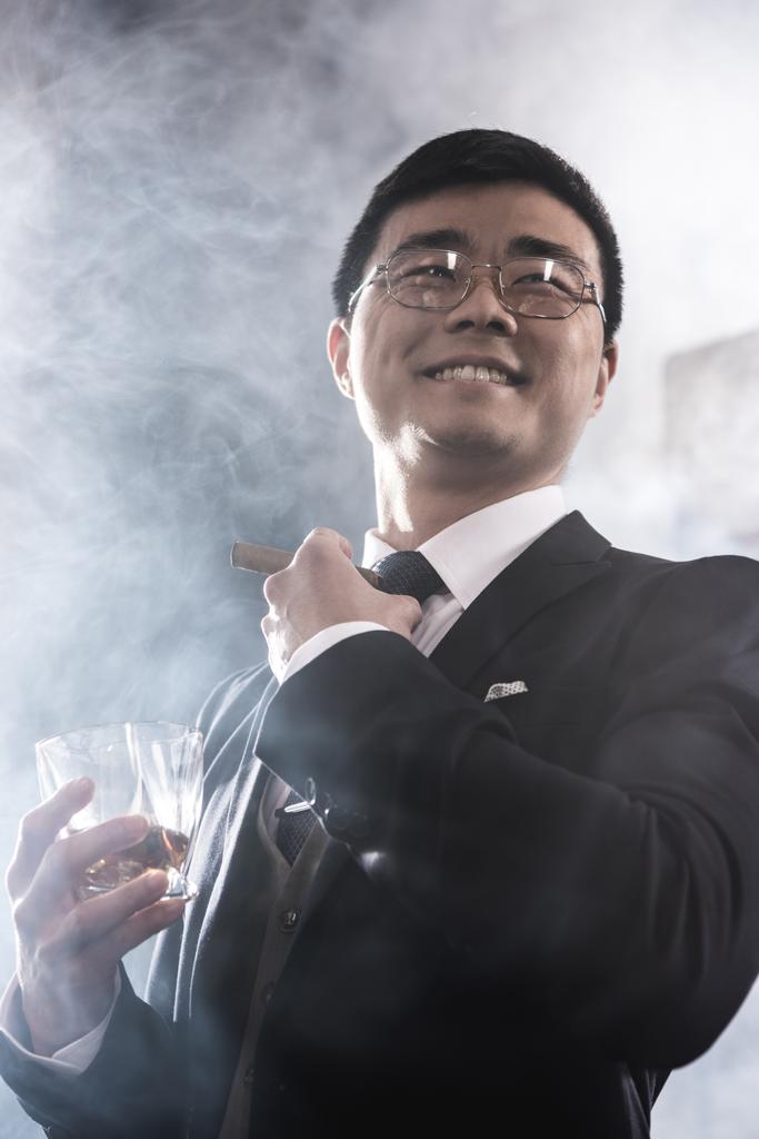 Nízký úhel pohledu úsměvu asijské muž v brýlích pít whisky a kouřit doutník - Fotografie, Obrázek
