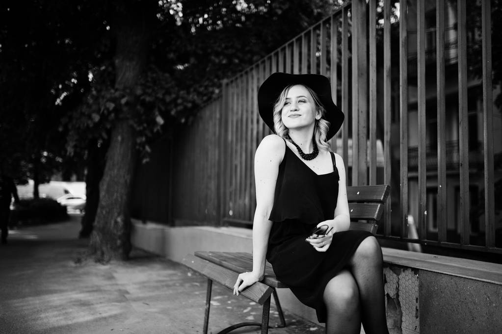 Blondynka na czarnej sukni, naszyjniki i kapelusz, siedząc na ławce. - Zdjęcie, obraz