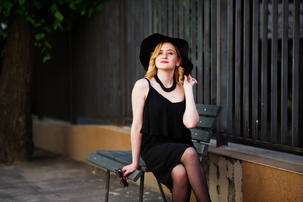 黒のドレス、ネックレス、帽子のベンチに座っている金髪の女性. - 写真・画像
