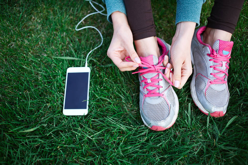 urheilullinen nainen urheiluvaatteissa istuu älypuhelimen kanssa nurmikolla puistossa ja sitoo kengännauhat lenkkarit
 - Valokuva, kuva