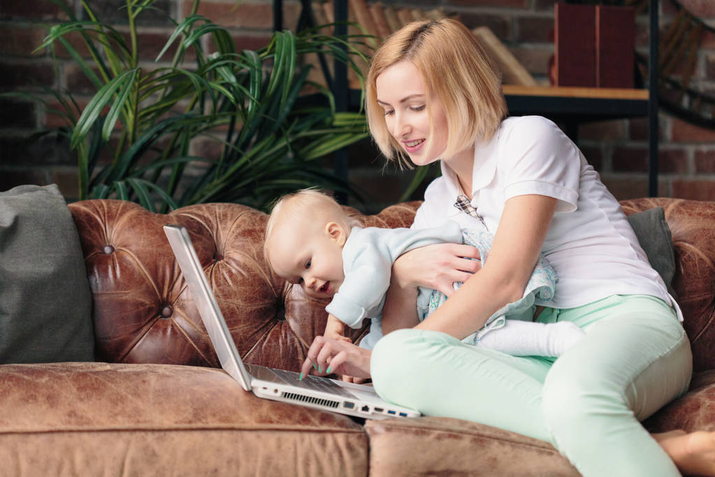 Mãe sentada no sofá enquanto trabalhava no laptop com um bebê
 - Foto, Imagem