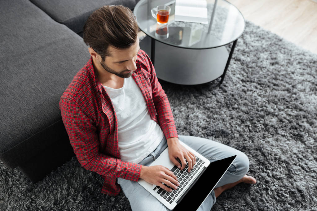 Νεαρός άνδρας κάθεται σε κλειστούς χώρους στο σπίτι ενώ χρησιμοποιείτε το φορητό υπολογιστή - Φωτογραφία, εικόνα
