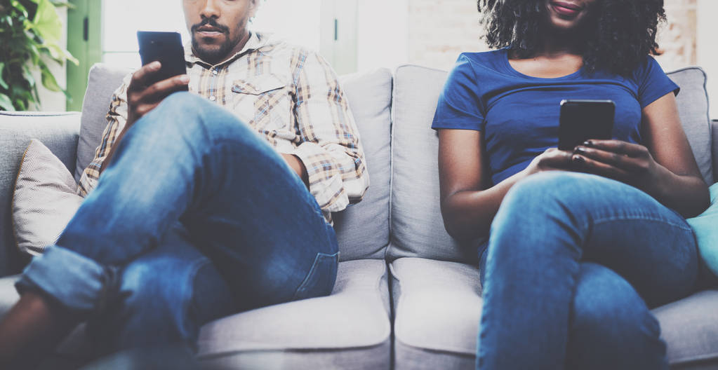 Грустная африканская американская пара расслабляется вместе на sofa.Young черный человек и его подруга, используя мобильные устройства во время отдыха дома в гостиной. Horizontal, blurred background.Cropped
 - Фото, изображение