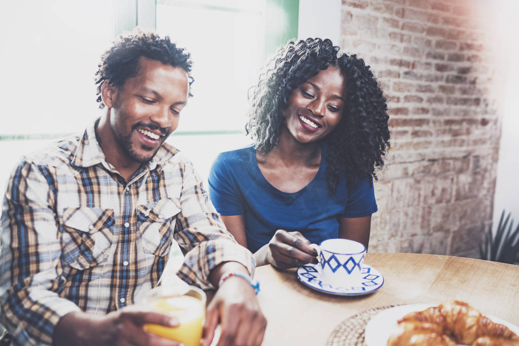 Felice coppia afro-americana stanno facendo colazione insieme al mattino al tavolo di legno. sorridente uomo nero e la sua ragazza bere succo di frutta fresco e caffè nero a colazione a casa
. - Foto, immagini
