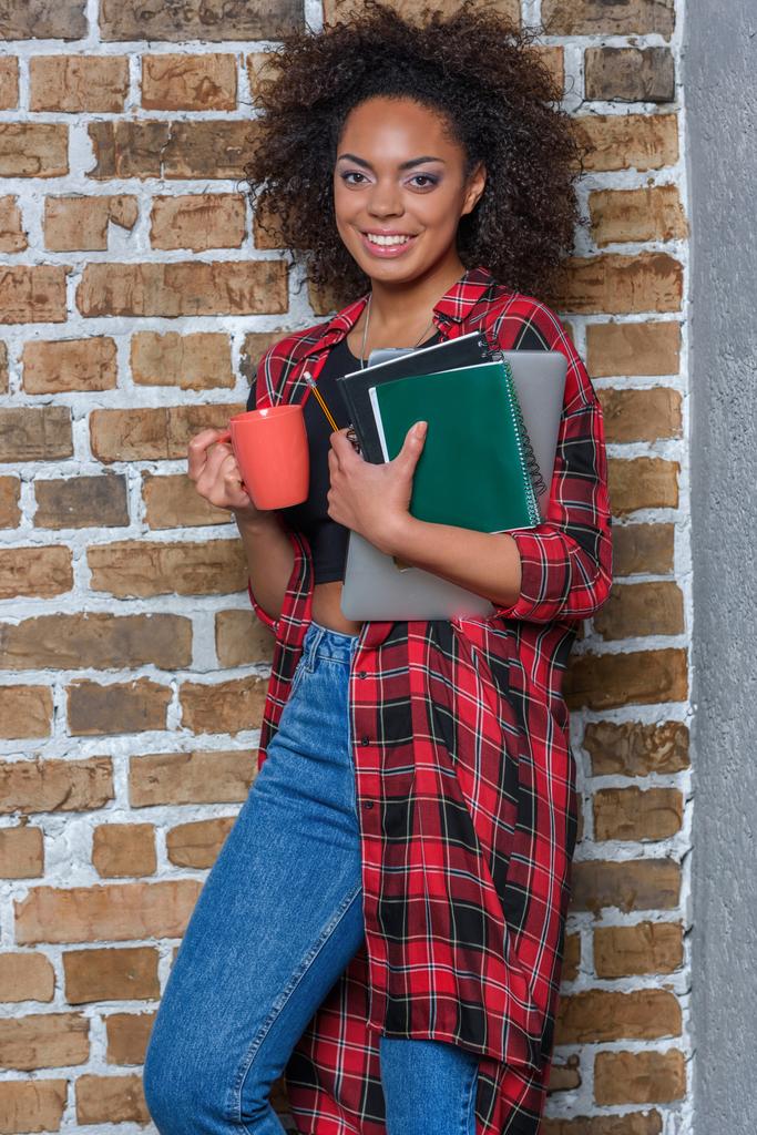 junge stylische Afroamerikanerin trinkt Kaffee und hält Notizbuch und Laptop in der Hand - Foto, Bild