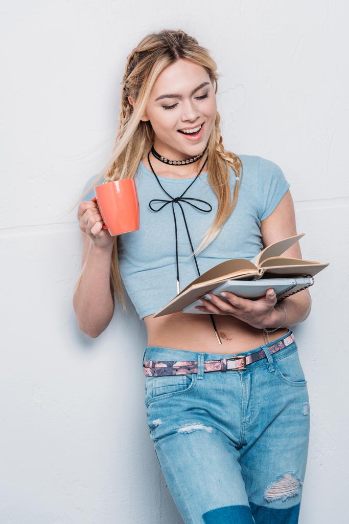 kaukasisches blondes Mädchen trinkt Kaffee und liest Bücher mit Gesichtsausdruck - Foto, Bild