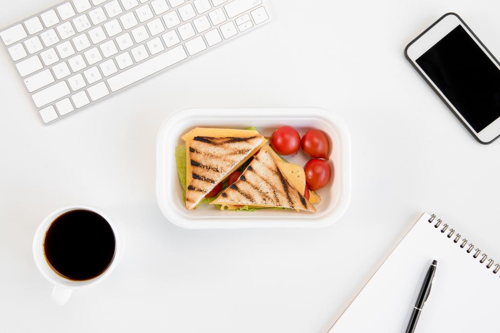 Το Top view σάντουιτς με ντομάτες στο καλαθάκι, σημειωματάριο με στυλό, φλιτζάνι καφέ, smartphone και πληκτρολόγιο στο χώρο εργασίας - Φωτογραφία, εικόνα