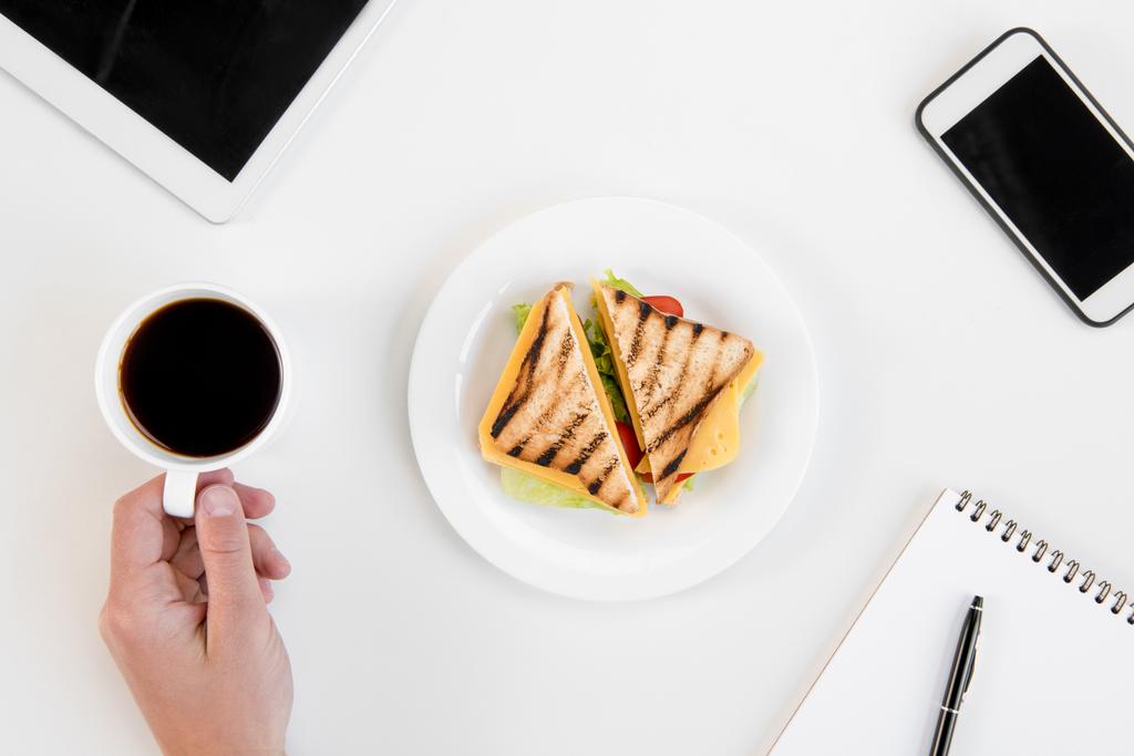 Vista superior de la persona comiendo sándwiches y tomando café en el lugar de trabajo con electrónica
  - Foto, Imagen