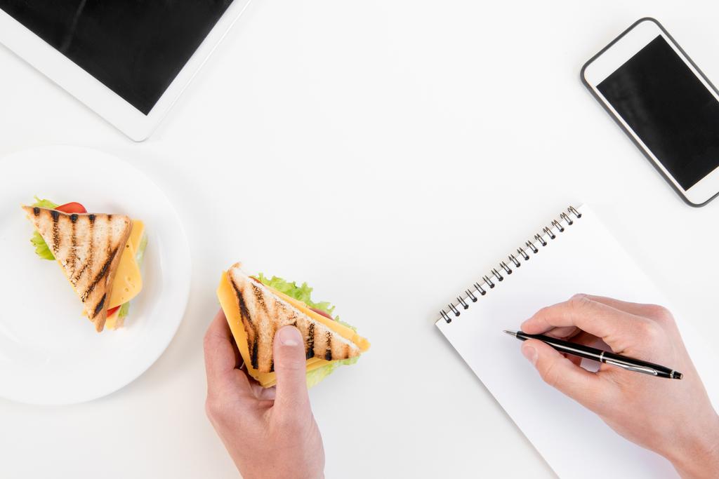 Ansicht von oben: Person schreibt in Notizbuch, während sie am Arbeitsplatz Sandwiches isst - Foto, Bild