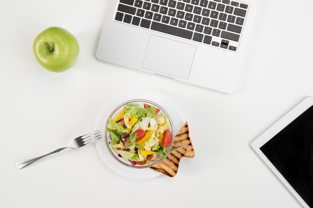 Vista superior del portátil y la tableta digital con pantalla en blanco, manzana verde fresca y ensalada fresca con tostadas en el lugar de trabajo
 - Foto, imagen
