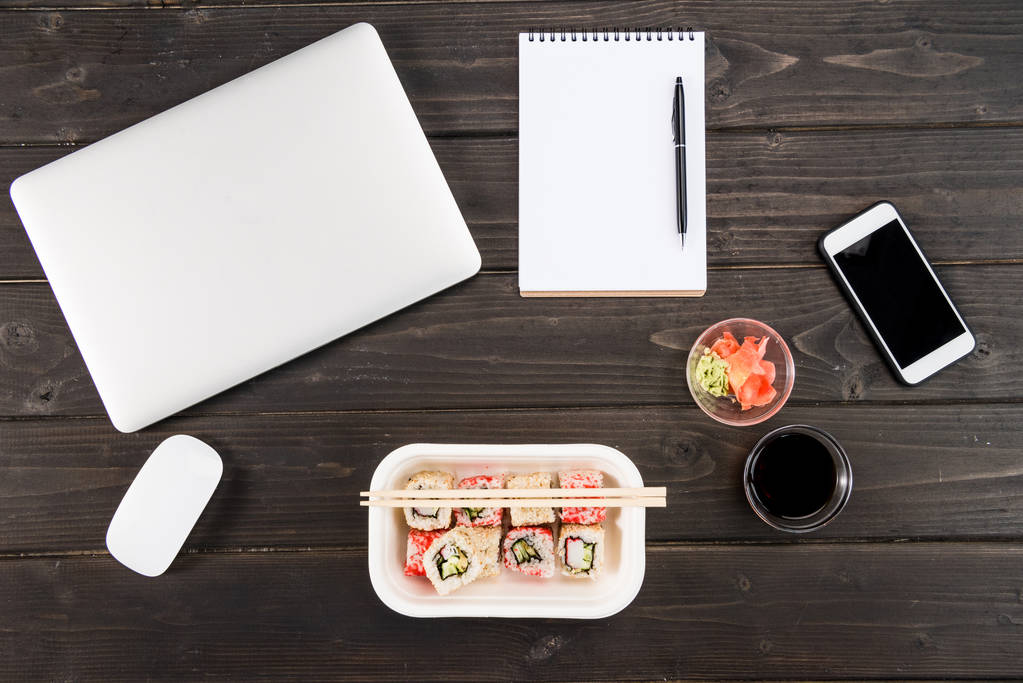 Вид сверху ноутбука с компьютерной мышью, блокнота с ручкой, смартфоном и набором суши на деревянном столе
  - Фото, изображение