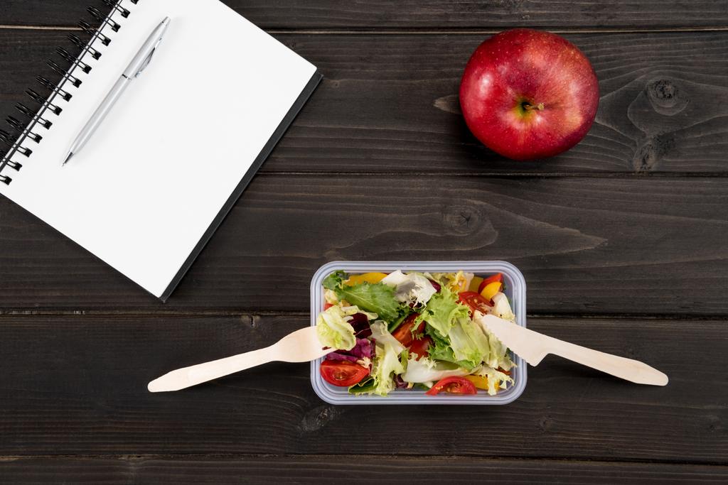 Top view of natural salad with red apple and notebook on wooden surface - Photo, Image