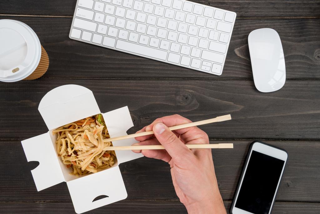 hand holding chopsticks with noodle and keyboard with smartphone on wooden tabletop - Photo, Image