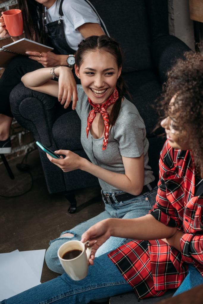 Jovens mulheres atraentes com smartphone e xícara de café sorrindo umas às outras
 - Foto, Imagem
