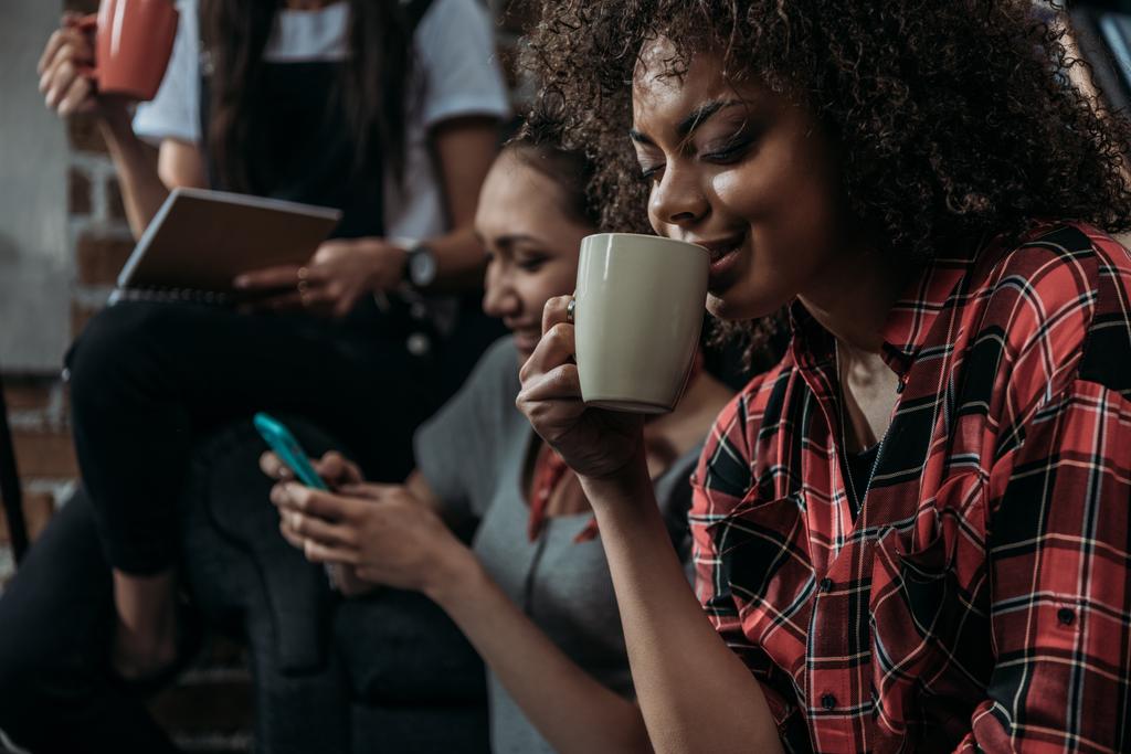 Onnellinen nuori afrikkalainen amerikkalainen nainen juo kahvia, kun ystävät käyttävät älypuhelinta ja pitävät kuppia takana
 - Valokuva, kuva