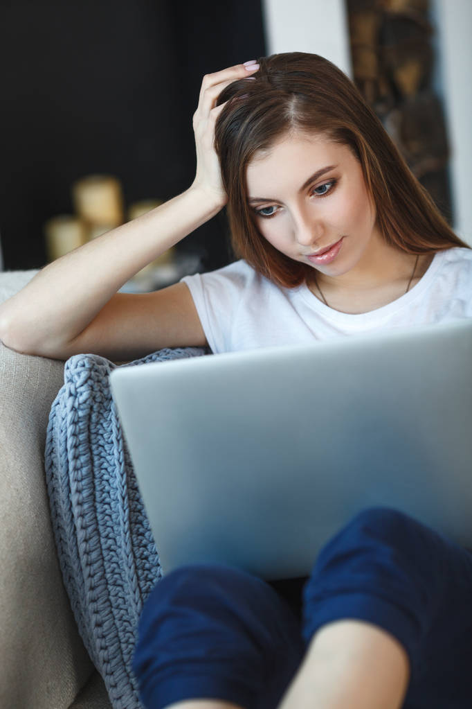 Nuori nainen istuu kannettavan tietokoneen kanssa sohvalla kotonaan
 - Valokuva, kuva