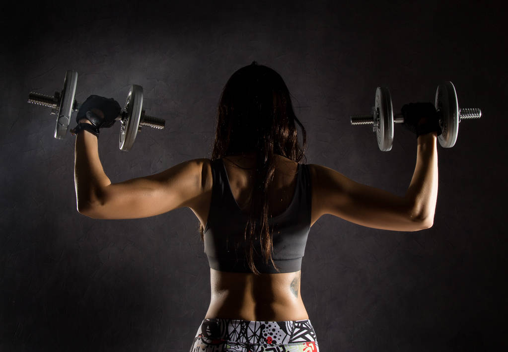 back view of sporty sexy girl with dumbbells on a dark background. Athlete doing exercises in the gym - Photo, Image