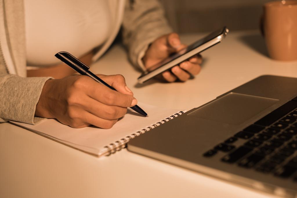 γυναίκα γράφοντας στο σημειωματάριο ενώ κρατάτε το smartphone - Φωτογραφία, εικόνα