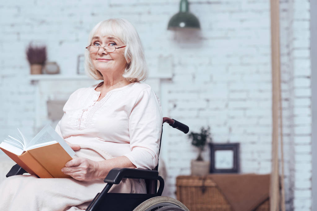 Oude vrouw lezen boek in rolstoel thuis uitgeschakeld - Foto, afbeelding