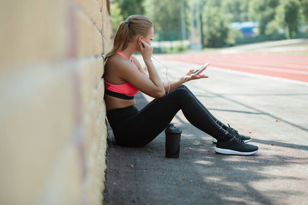 Νεαρή γυναίκα σπορ αθλητής σε αθλητικά είδη συνεδρίαση, ακούτε μουσική με ακουστικά και να χαλαρώσετε στο στίβο του σταδίου μετά από σκληρή προπόνηση - Φωτογραφία, εικόνα