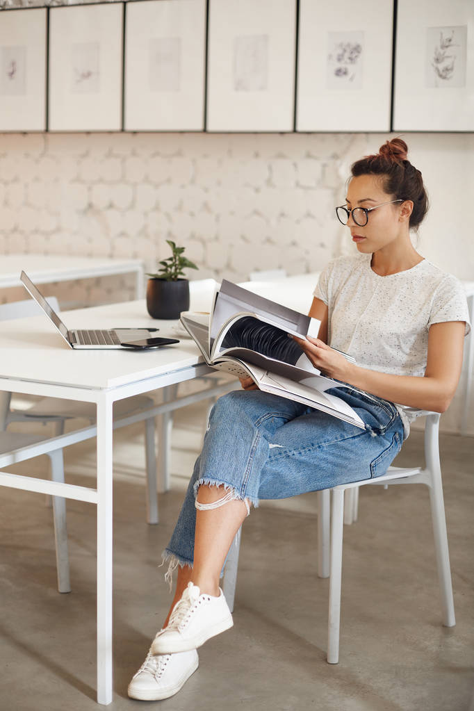 Femme dans la vingtaine poursuivant une carrière dans le design de mode en regardant un magazine assis dans son studio lumineux avec ordinateur portable
. - Photo, image