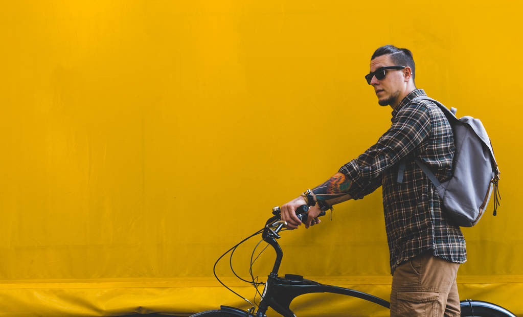 Hombre adulto guapo en gafas de sol camina con una bicicleta a lo largo de una pared amarilla Copiar espacio
 - Foto, Imagen