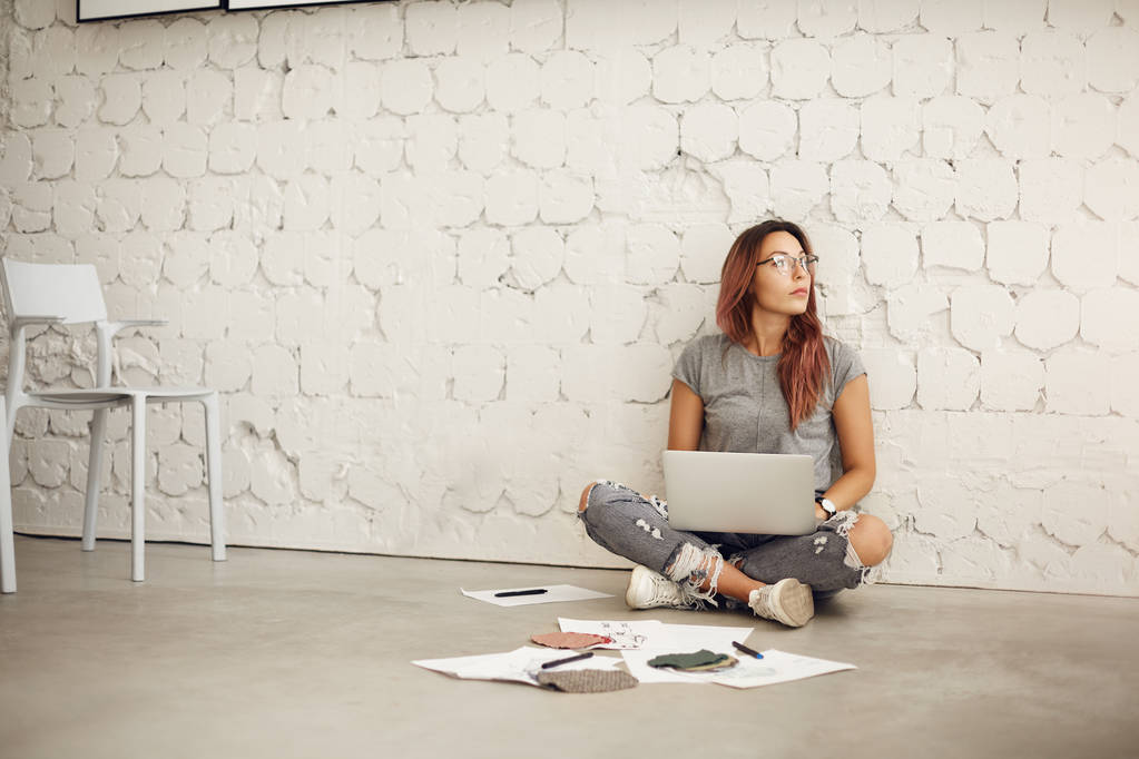 Weitwinkelaufnahme einer kreativen Mitarbeiterin, die mit Laptop auf dem Boden sitzt und eine Pause von ihrem täglichen Job in einem hellen Atelierraum einlegt - Foto, Bild