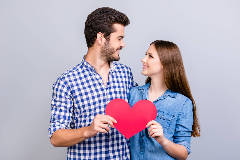 Love story. Trust and feelings, emotions and joy. Happy young lovely couple in love is posing, wearing casual shirts, holding big red paper heart and smiling on the pure background - Photo, Image