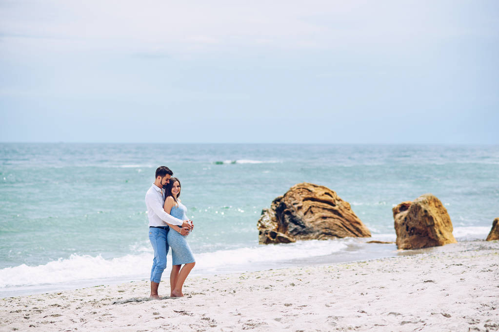 Homem corajoso e bonito em uma camisa branca e calças azuis abraça sua bela esposa grávida em um vestido azul contra o fundo das rochas e do mar
. - Foto, Imagem
