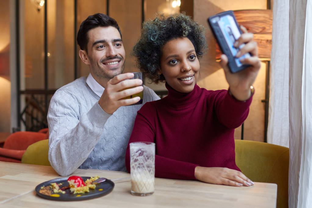 Szczęśliwy międzyrasowy pary widuje się w kawiarni w godzinach popołudniowych, ciemnoskóry kobieta jest rozciąganie jej ręki trzymającej telefon, aby zrobić zdjęcie jej i chłopak, oba są uśmiechający się pozytywnie i pokazywanie zawartości. - Zdjęcie, obraz