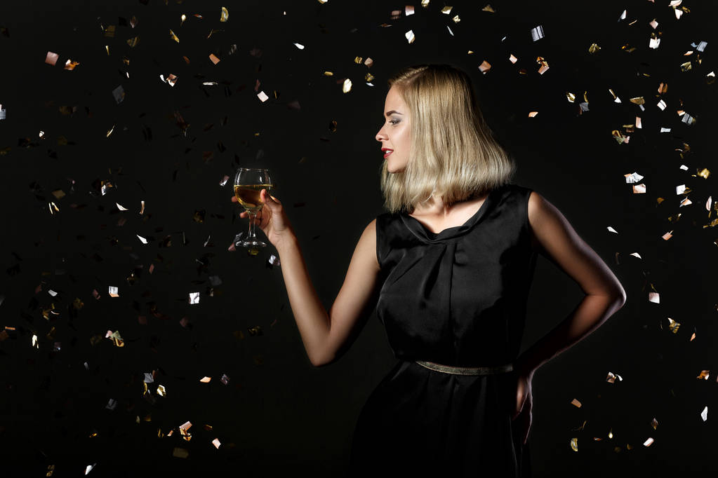 Kaunis onnellinen blondi nainen tilalla lasi valkoviiniä mustalla taustalla konfetti
 - Valokuva, kuva