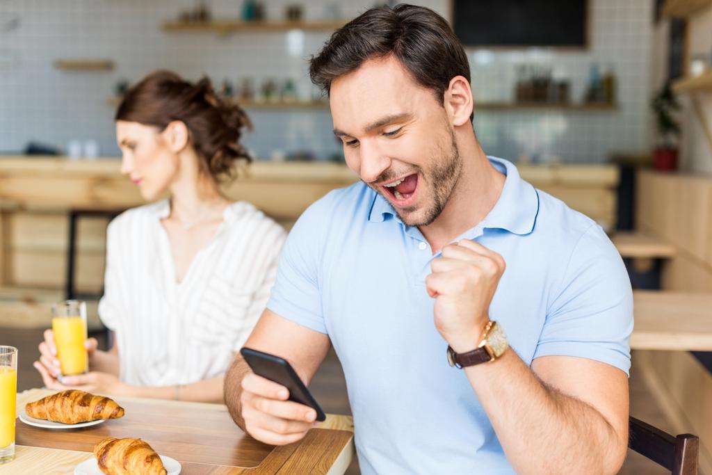 Ein süchtiger Mann mit seinem Smartphone und eine unglückliche Frau, die im Hintergrund sitzt. Mann feiert Gewinn einer Wette mit einer Glücksspiel-Handy-App. Fußballwetten, Sportwetten - Foto, Bild