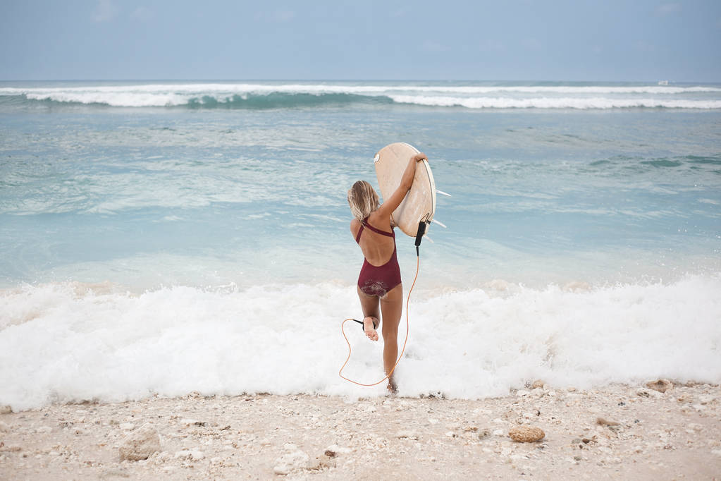 Retrato panorâmico horizontal de uma menina de surf atraente em terno de natação vermelho escuro está indo para o oceano com uma prancha de surf, vista de trás
 - Foto, Imagem