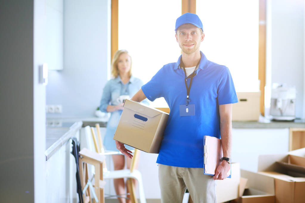 Lachende bezorger in blauw uniform levert pakketdoos aan ontvanger - koerier service concept. Glimlachende bezorger in blauw uniform - Foto, afbeelding