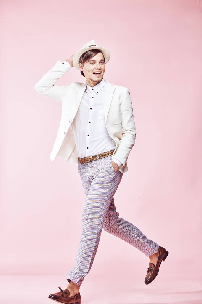 Jovem bonito feliz sorrindo homem moderno vestindo jaqueta branca, camisa branca, calças cinza e chapéu branco posando em estúdio com fundo rosa claro e segurando a mão no chapéu
 - Foto, Imagem