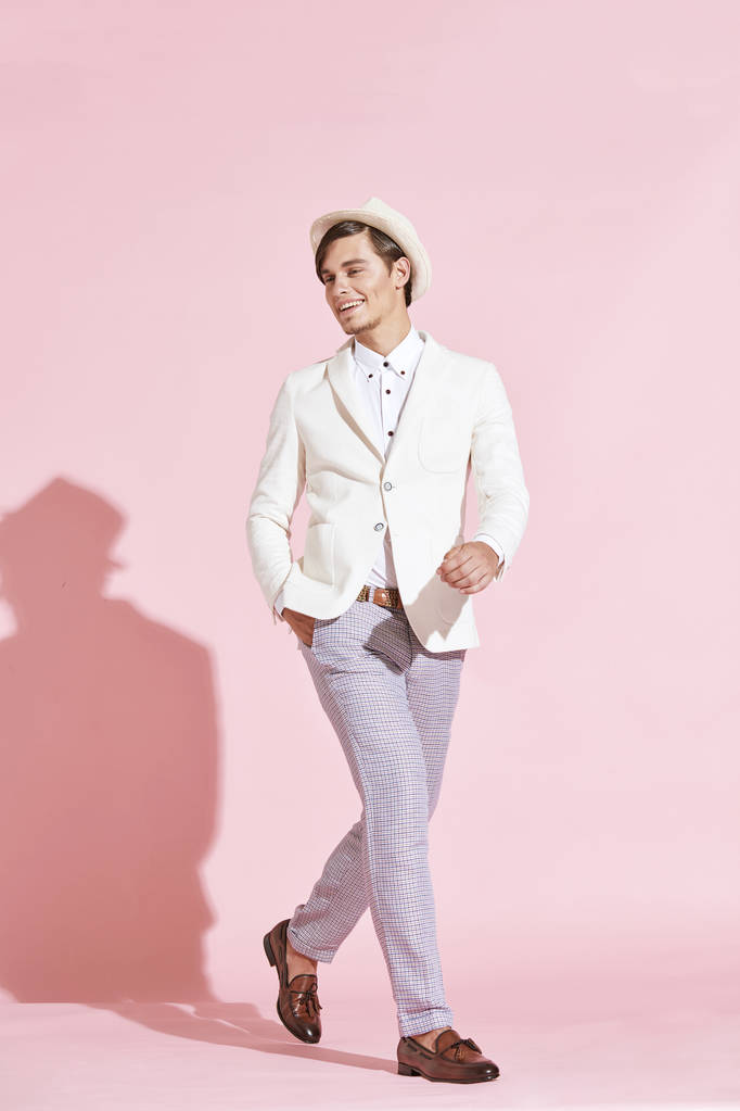 Gyönyörű komoly modern fiatalember visel fehér kabátot, fehér inget, szürke nadrágot és pózol stúdió világos rózsaszín háttér, fehér kalapot - Fotó, kép