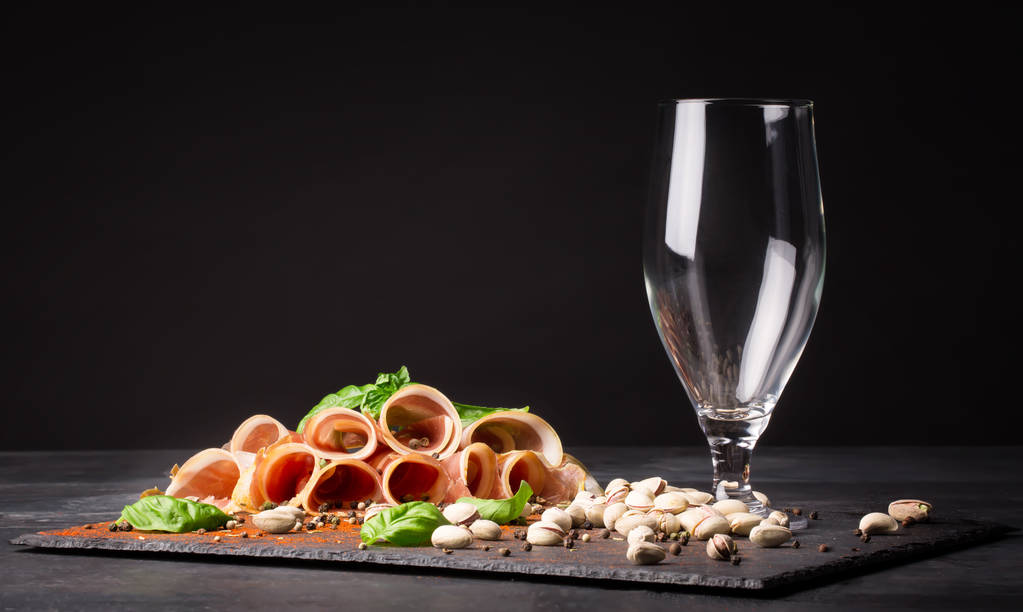 An empty glass of beer with pistachios and prosciutto seasoned with black pepper on a dark background. - Photo, Image
