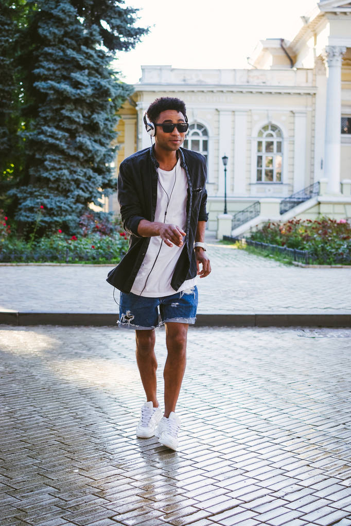 Portrait of young stylish hipster black man in white headphones and sun glasses dancing outdoor in city centre, having some fun - Photo, Image