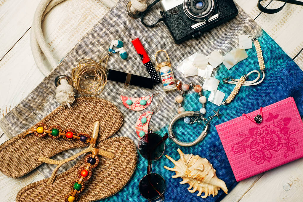  Επίπεδη θέσει καλοκαίρι μόδας με κάμερα, παντόφλες, γυαλιά ηλίου και άλλα αξεσουάρ κορίτσι πάνω από την τσάντα σε άσπρο φόντο ξύλινη - Φωτογραφία, εικόνα