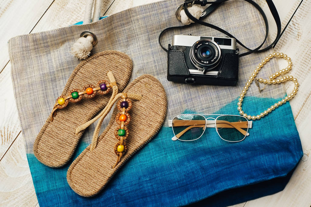  Lapos feküdt a nyári divat kamera, papucs, napszemüveg és egyéb lány kiegészítők tetején a zsák, fehér fa háttér - Fotó, kép