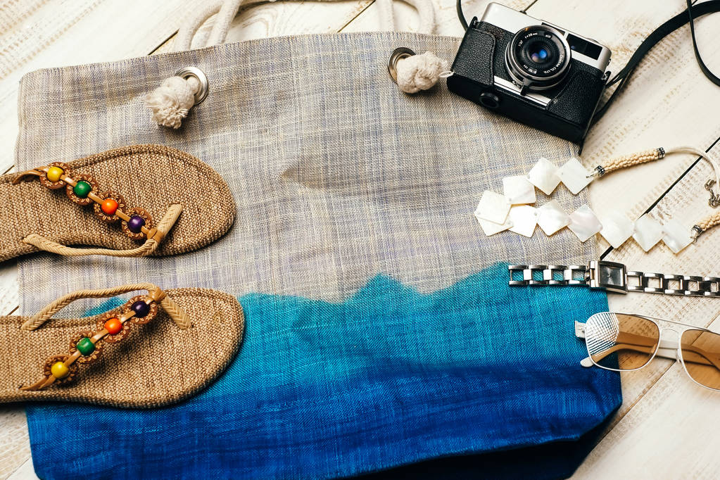  Ajuste plano de la moda de verano con cámara, zapatillas, gafas de sol y otros accesorios de niña en la parte superior del bolso sobre fondo de madera blanca
 - Foto, imagen