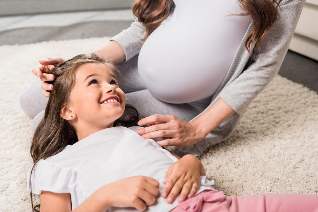 Беременная женщина с дочерью, сидящей на ковре
  - Фото, изображение