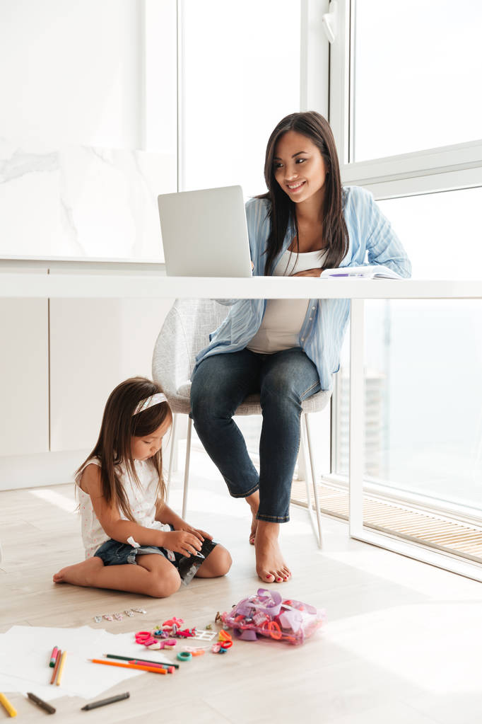 Koncentrált nő használ laptop lánya padlón ül míg. - Fotó, kép