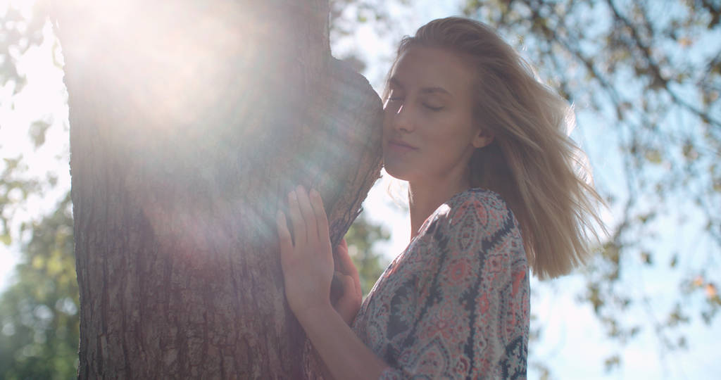 Νεαρή φυσικά όμορφη γυναίκα αγκαλιάζει ένα δέντρο στο ηλιόλουστο πάρκο. - Φωτογραφία, εικόνα