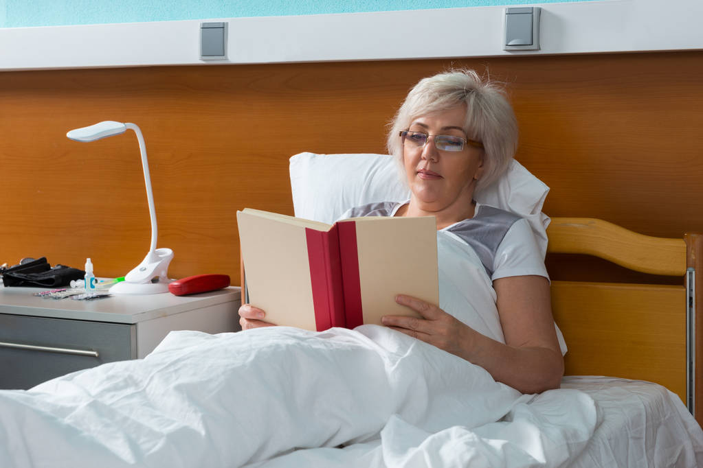 女性患者は病院のベッドで横たわっている間、本を読んでいます。 - 写真・画像